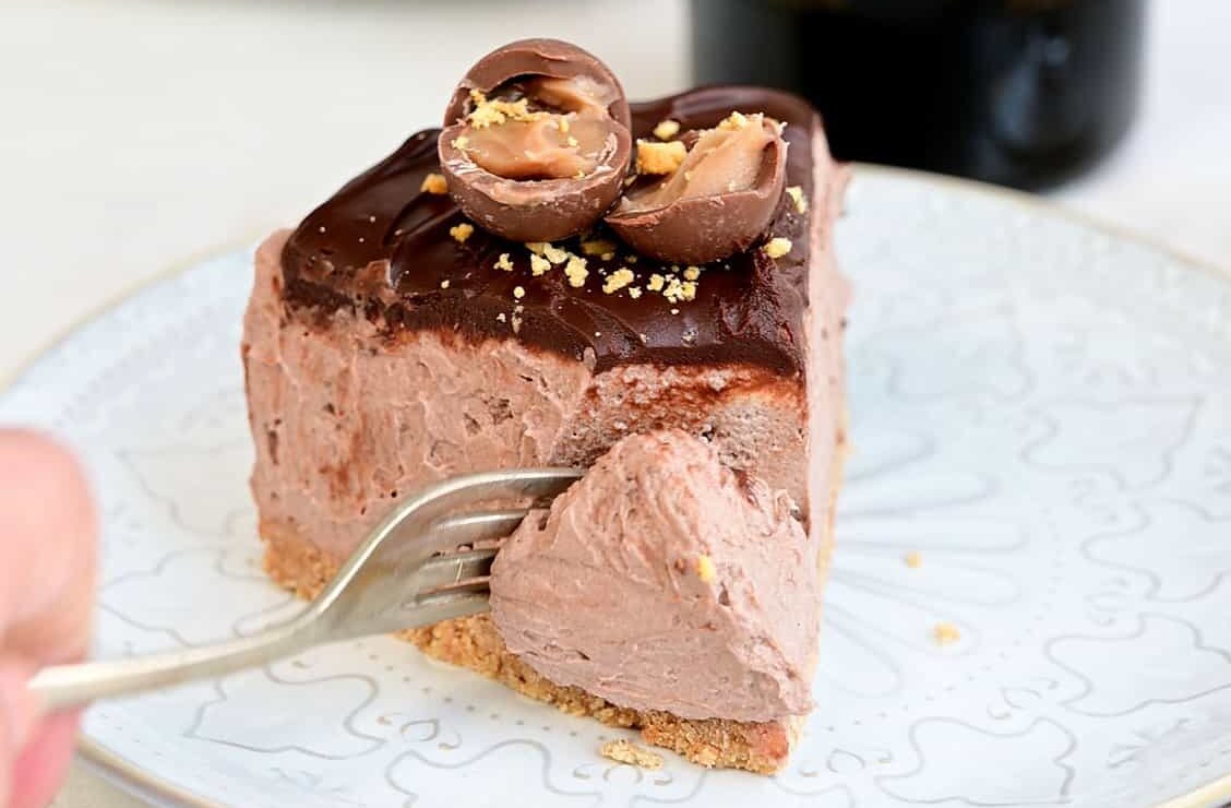 A slice of no bake Baileys Cheesecake topped with two chocolate truffles and chocolate sauce sits on a plate, as a fork cuts in. In the background are stacked plates and a bottle of Baileys Irish Cream, adding to the indulgent scene.