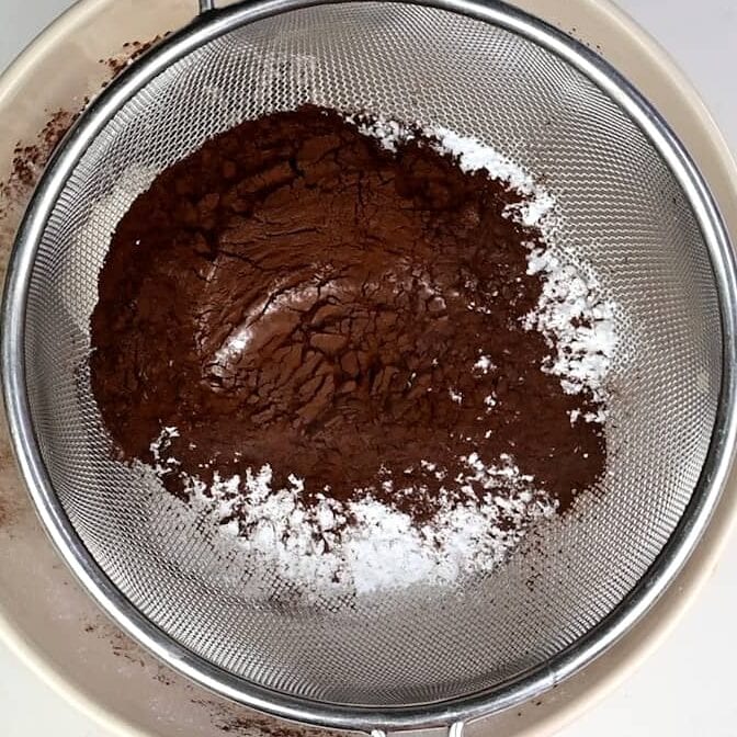 A metal sieve holds a mixture of cocoa powder and powdered sugar above a white bowl, ready to be sifted for baking a delicious no bake Baileys Cheesecake.