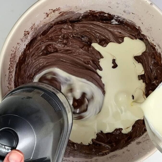 A hand holds an electric mixer blending chocolate batter in a bowl while another pours in creamy liquid, likely Baileys, perfect for a no bake Baileys cheesecake recipe.