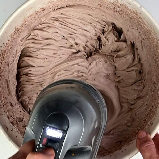 A hand holds an electric mixer, blending a large bowl of light brown, creamy no bake Baileys Cheesecake batter. Swirls and peaks are visible in the mixture.