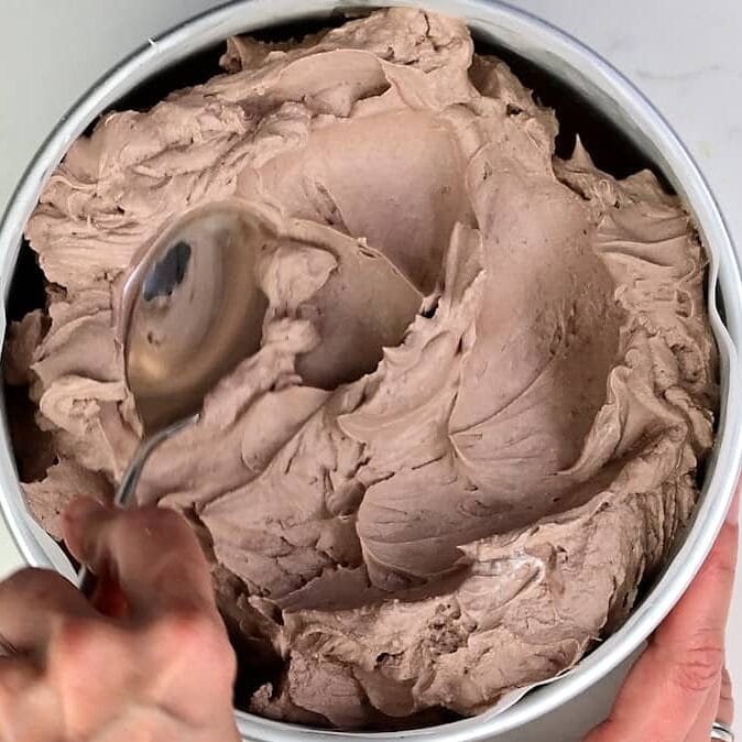 A hand uses a spoon to scoop creamy chocolate ice cream from a round container, viewed from above—perfect alongside a slice of no bake Baileys Cheesecake.