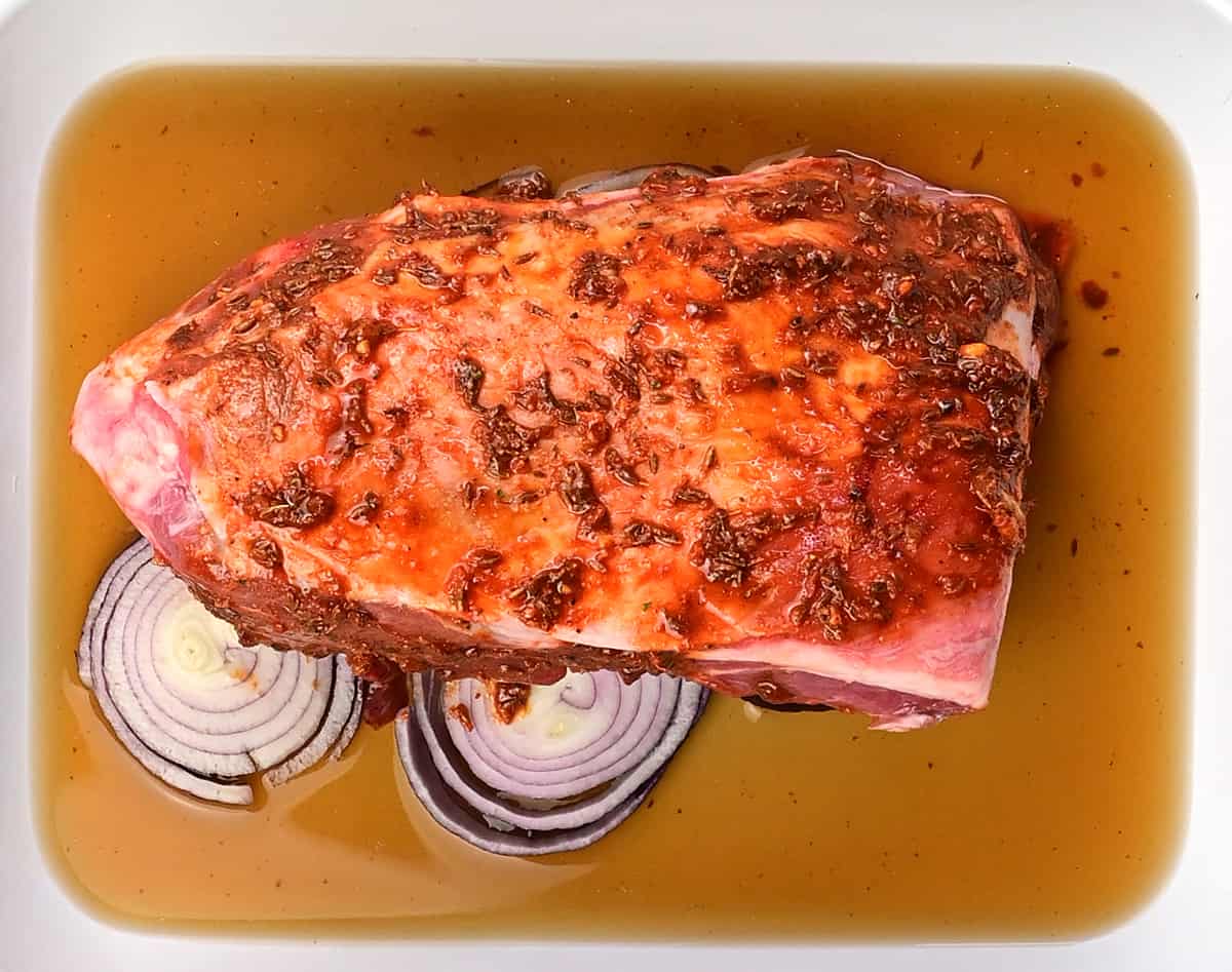 Raw lamb shoulder covered with marinade over sliced onions in a roasting tin