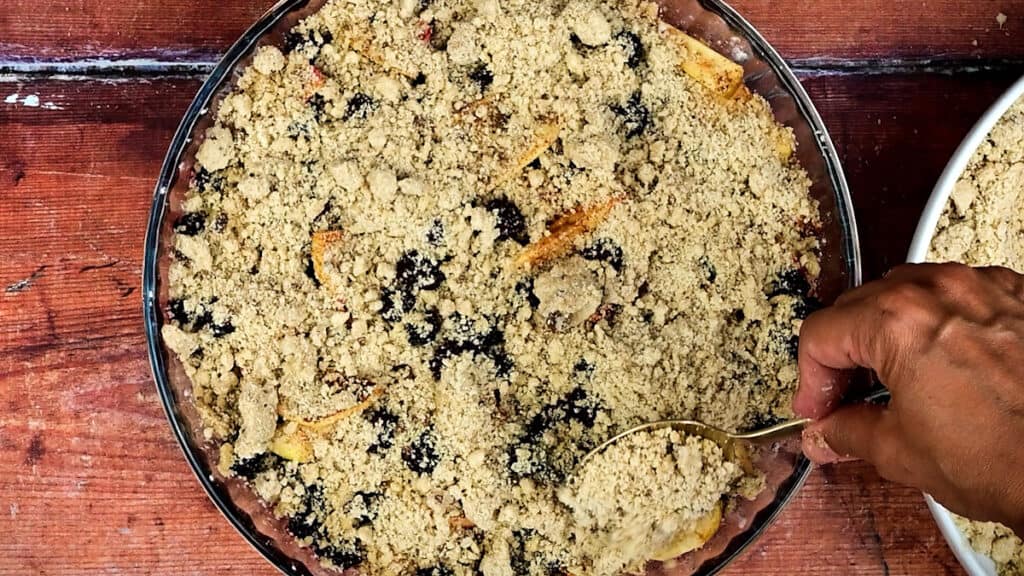 adding crumble topping over apples and blackberries in a glass pie dish