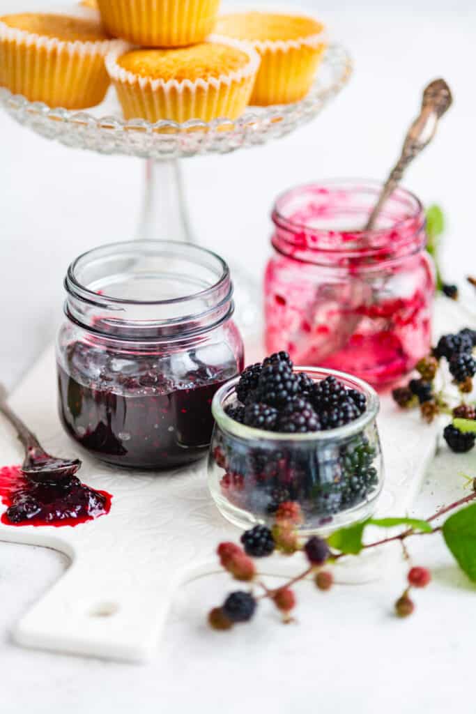 Jar of blackberry jam with small jar of wild blackberries on the side and stand of cupcakes in background