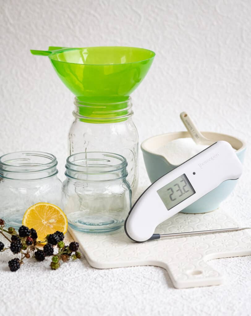 Ingredients and equipment for blackberry jam: jars, funnel, digital thermometer, blackberries, sugar, lemon