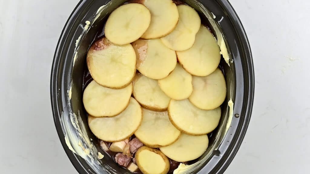 layering sliced potatoes over lamb stew ingredients in crockpot