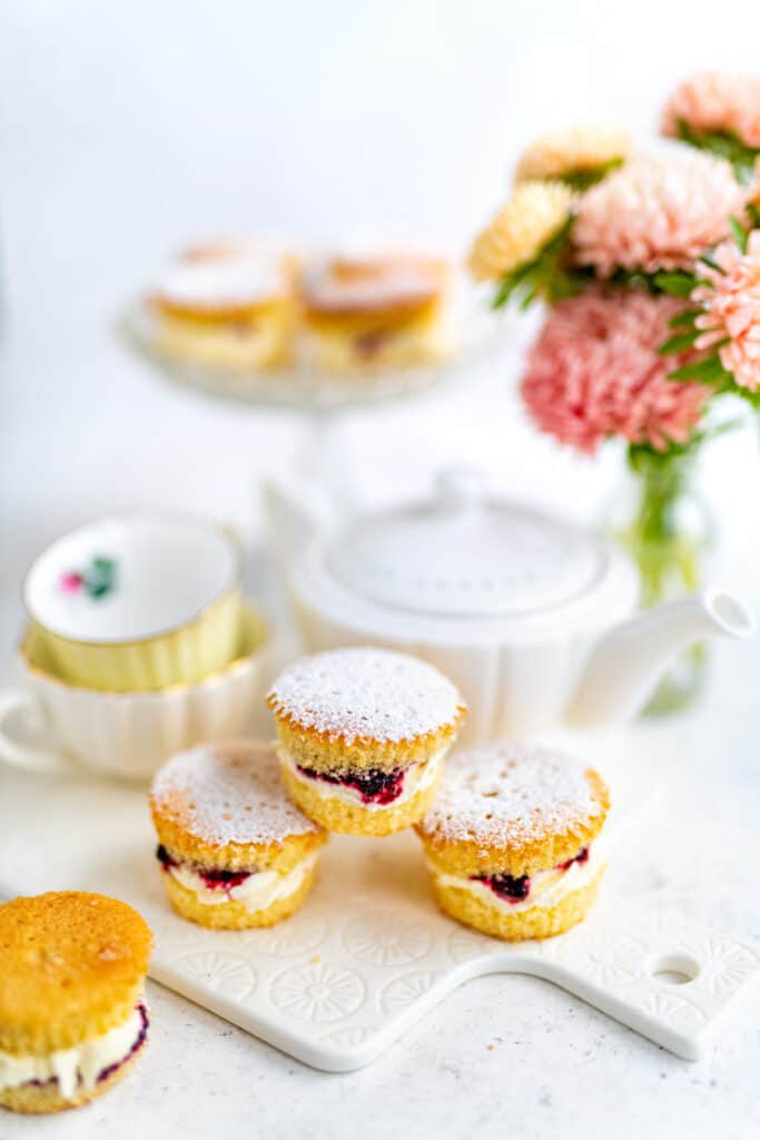 Victoria sponge cupcakes on a ceramic board with tea cups, tea pot and vase of flowers behind them