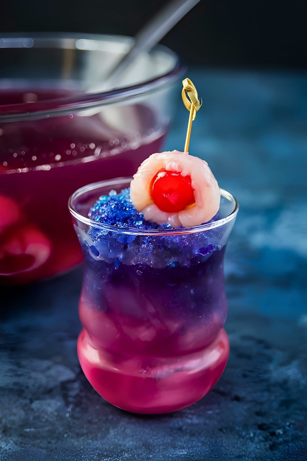 Purple People Eater Halloween cocktail with blue butterfly pea crushed ice melting into purple over a pink drink, topped with a lychee and cherry eyeball on a cocktail stick