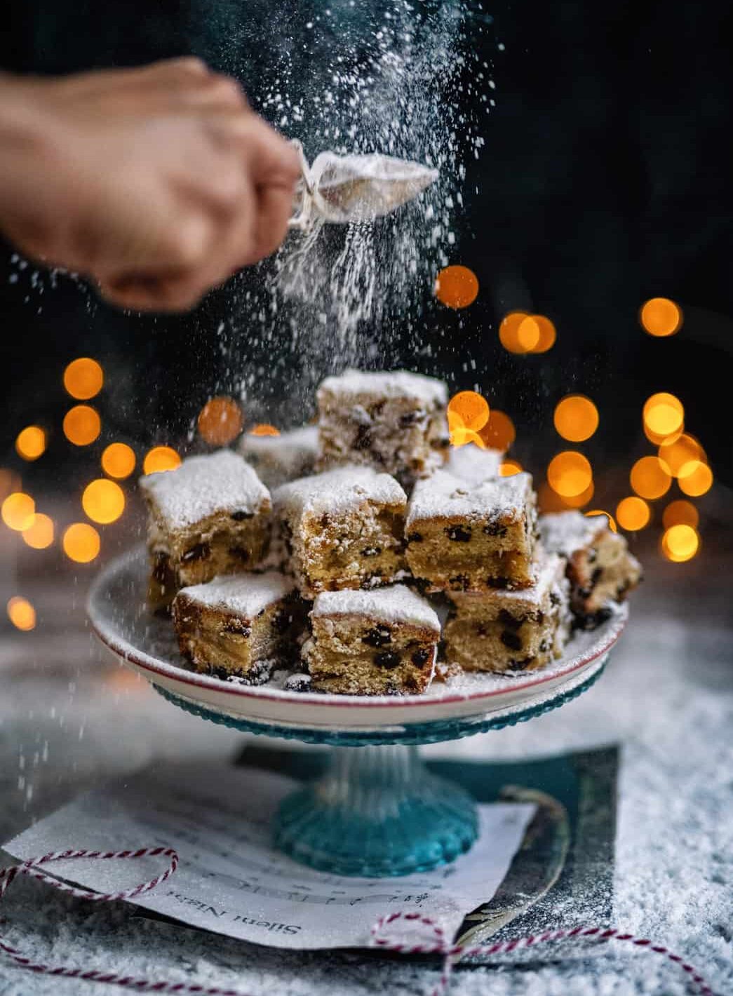 A hand sprinkles powdered sugar over a stack of German cake squares with fruit on a decorative cake stand. Warm, blurred lights glow in the background, creating a festive, cozy Christmas atmosphere&mdash;no yeast required.