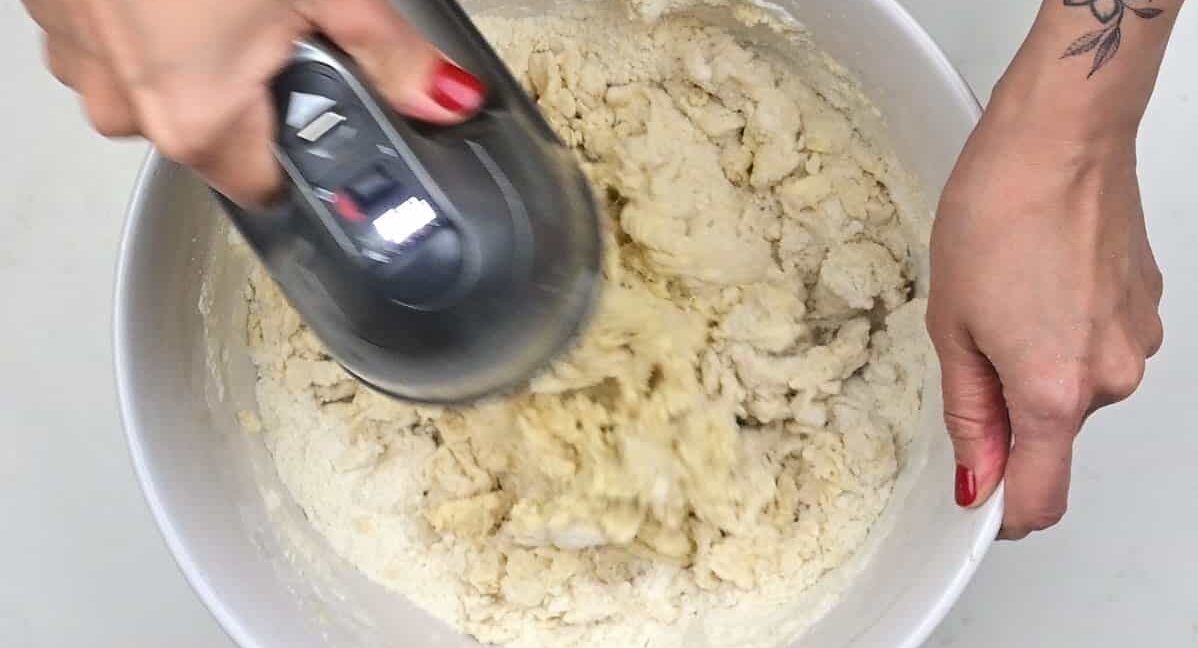 A person with red nails and a floral tattoo on their arm is mixing dough for stollen bites in a white bowl using a hand mixer.