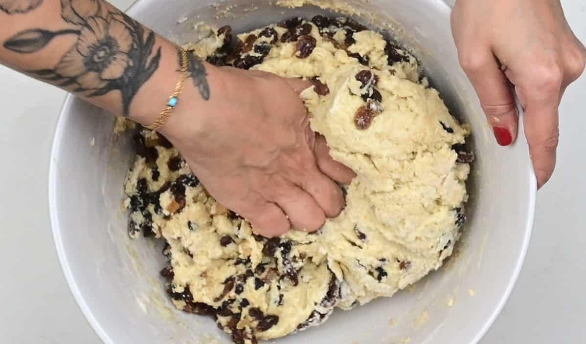 A person with a floral tattoo kneads dough with raisins for a stollen bites recipe in a large white mixing bowl, using their hands to mix the ingredients.