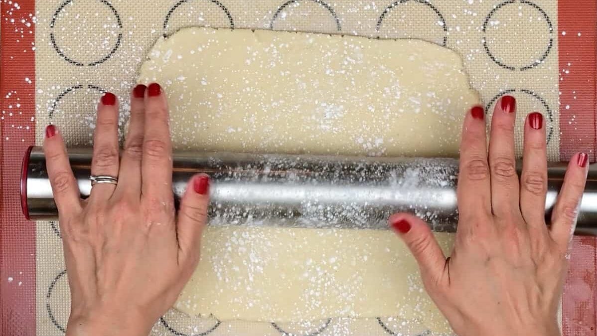 Hands with red nail polish use a metal rolling pin to roll out marzipan on a floured surface, preparing it for perfect Stollen Bites using circular outlines as a guide.