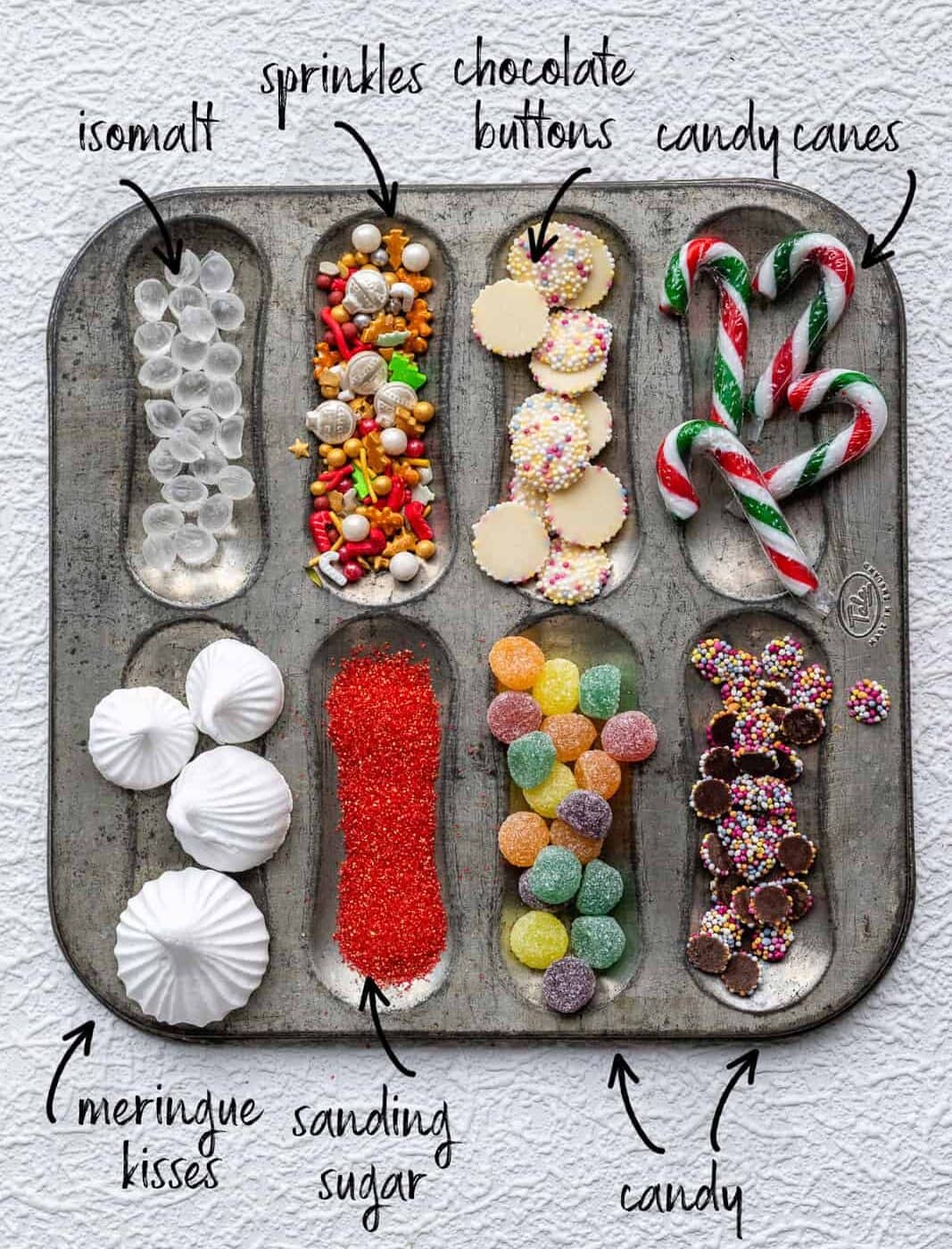 A muffin tin holds assorted holiday baking toppings: isomalt gems, colorful sprinkles, chocolate buttons, candy canes, meringue kisses, red sanding sugar, gumdrop candies—perfect for decorating with gingerbread house icing or your favorite Gingerbread House recipe.