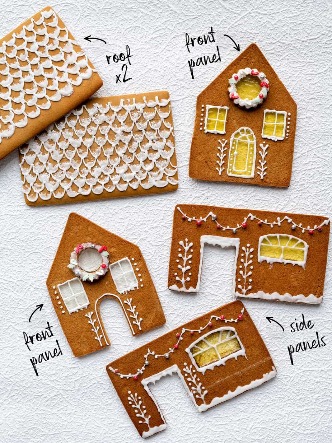 Five decorated Gingerbread House panels are arranged on a white surface: two roof pieces with scallop designs, a front panel with a wreath and window, another front panel, and two side panels detailed with gingerbread house icing.