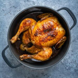 A Dutch oven roast chicken with golden brown, crispy skin sits in a black cast iron pot on a gray textured surface.