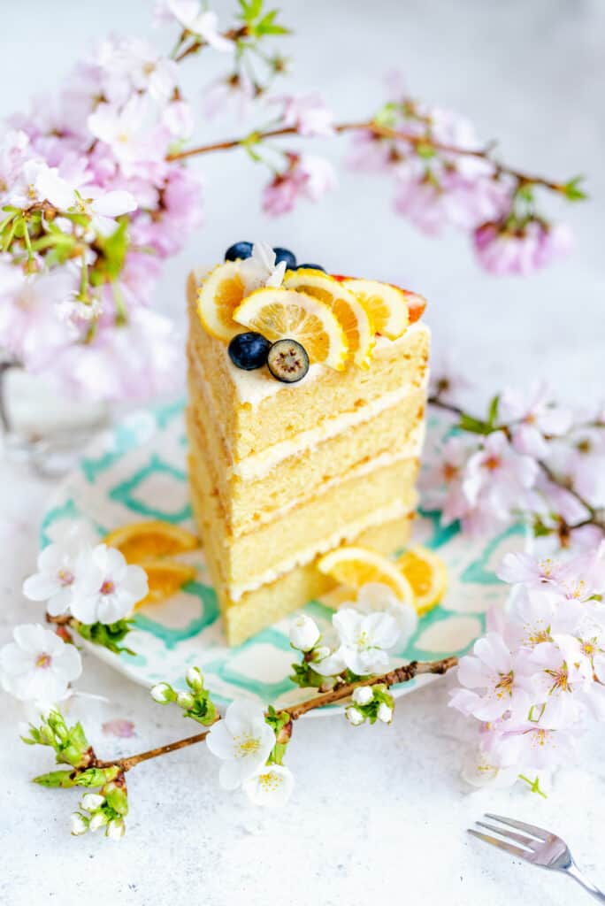 Slice of lemon cake decorated with lemon slices and blueberries among fresh flowers
