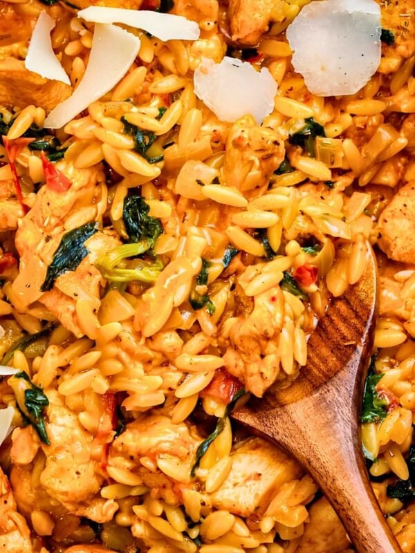 Close-up of creamy Cajun Chicken Orzo with chunks of chicken, spinach, tomato sauce, and parmesan cheese, as a wooden spoon scoops up the savory dish.