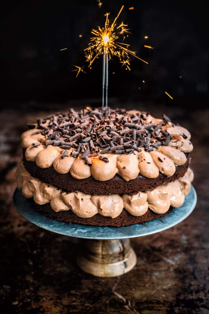 chocolate Victoria sandwich cake with chocolate frosting on a cake stand with a lighted sparkler