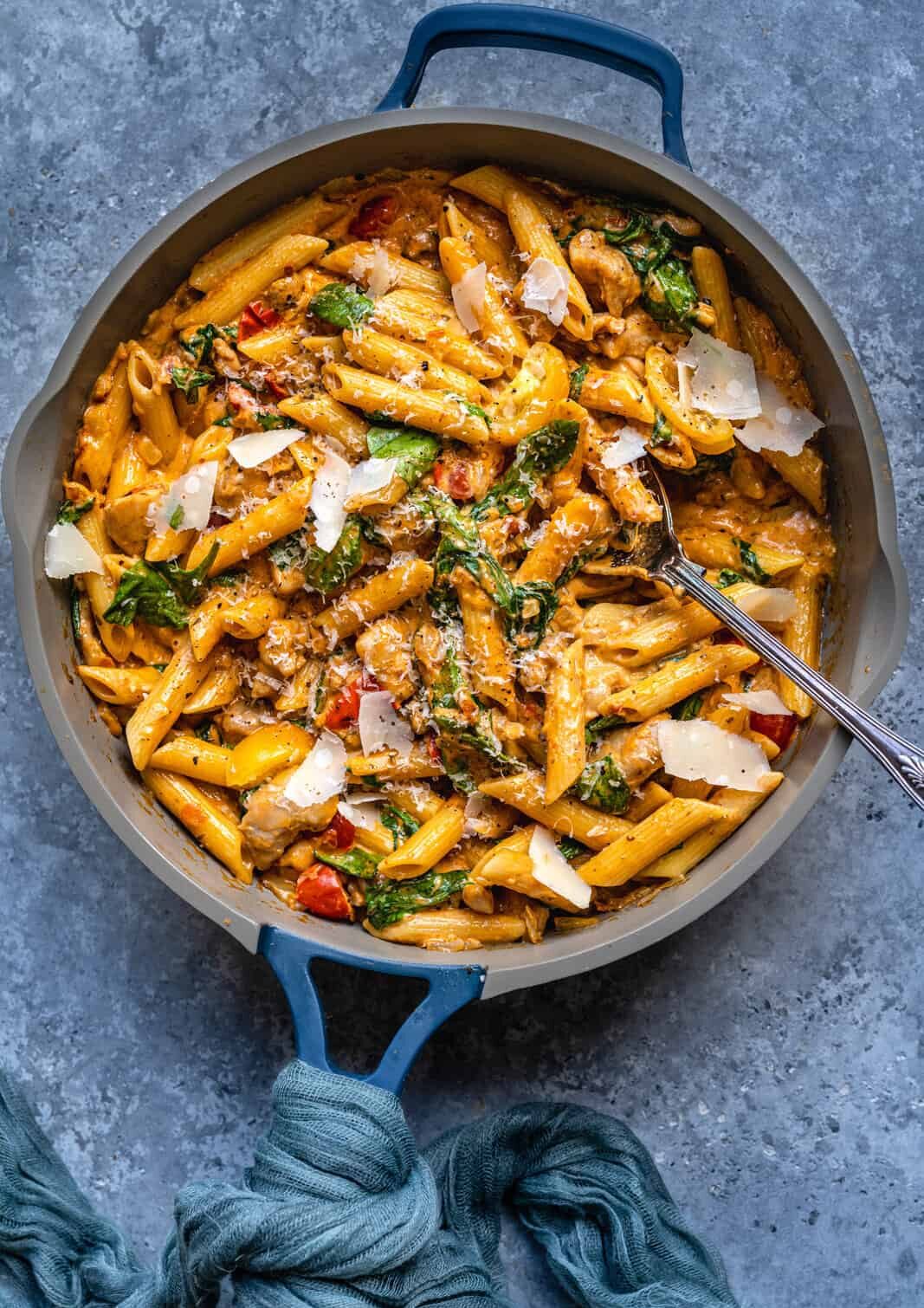 A skillet filled with creamy one pot tuscan chicken pasta, garnished with fresh spinach, cherry tomatoes, and shaved parmesan cheese. A serving spoon rests in the dish atop a gray surface with a blue cloth nearby.
