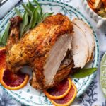 A roasted turkey breast with crispy skin is sliced and served on a decorative plate with blood orange slices and fresh sage, set on a patterned tablecloth next to a bowl of roasted root vegetables.