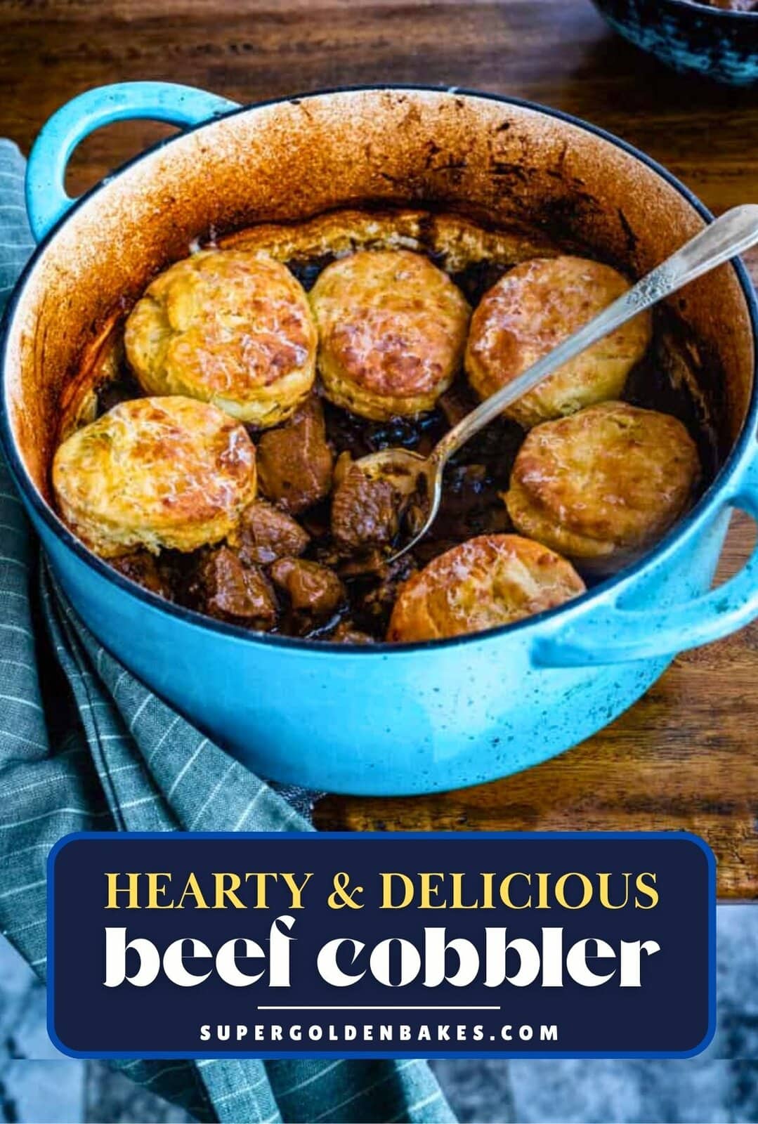A blue pot filled with hearty Beef Cobbler, topped with golden, flaky biscuits. A serving spoon rests inside, and a green cloth is draped nearby. Text on the image reads “Hearty & Delicious Beef Cobbler.”.