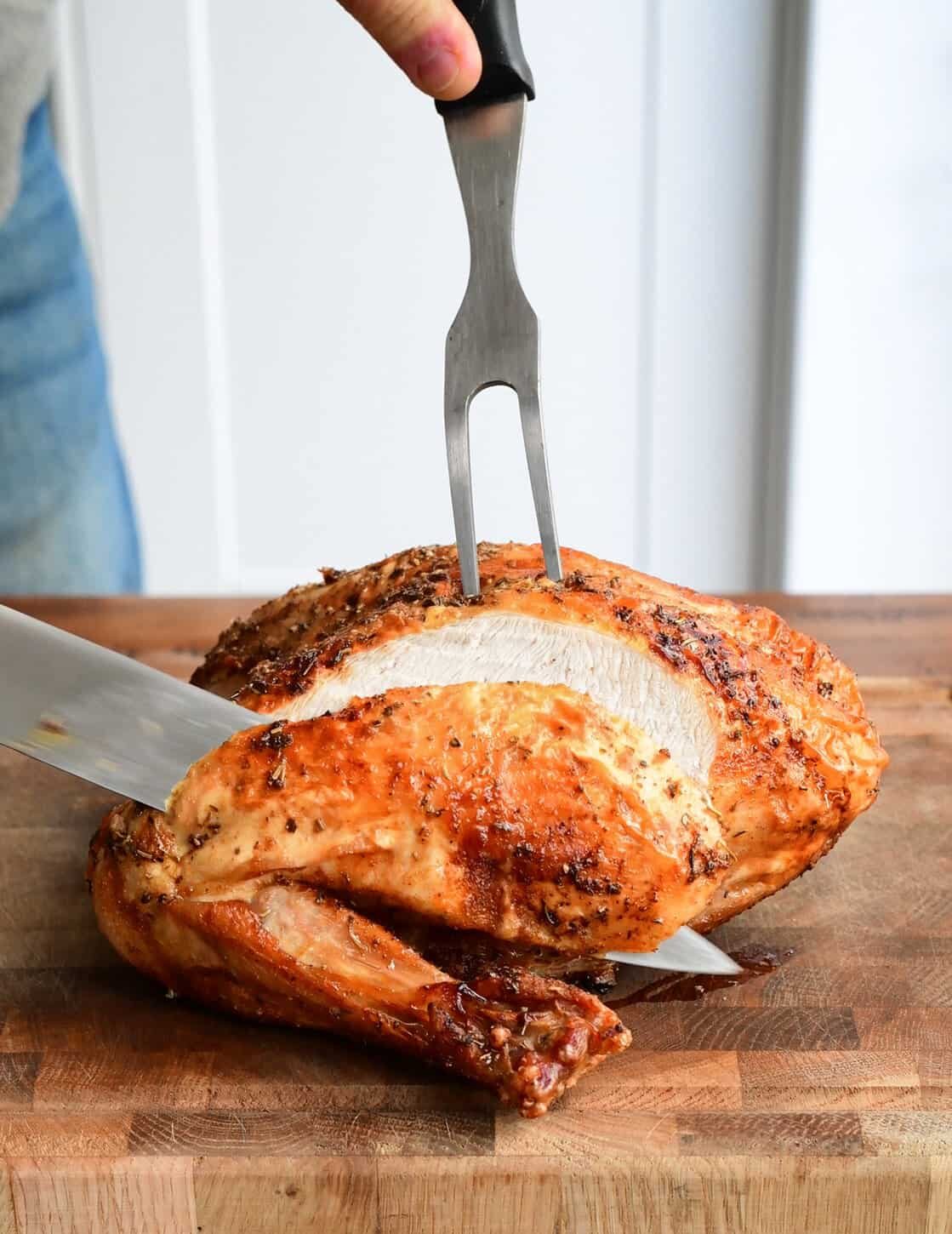 A person slices into a roasted air fryer turkey breast on a wooden cutting board using a large knife and a meat fork. The turkey skin is golden and crispy.