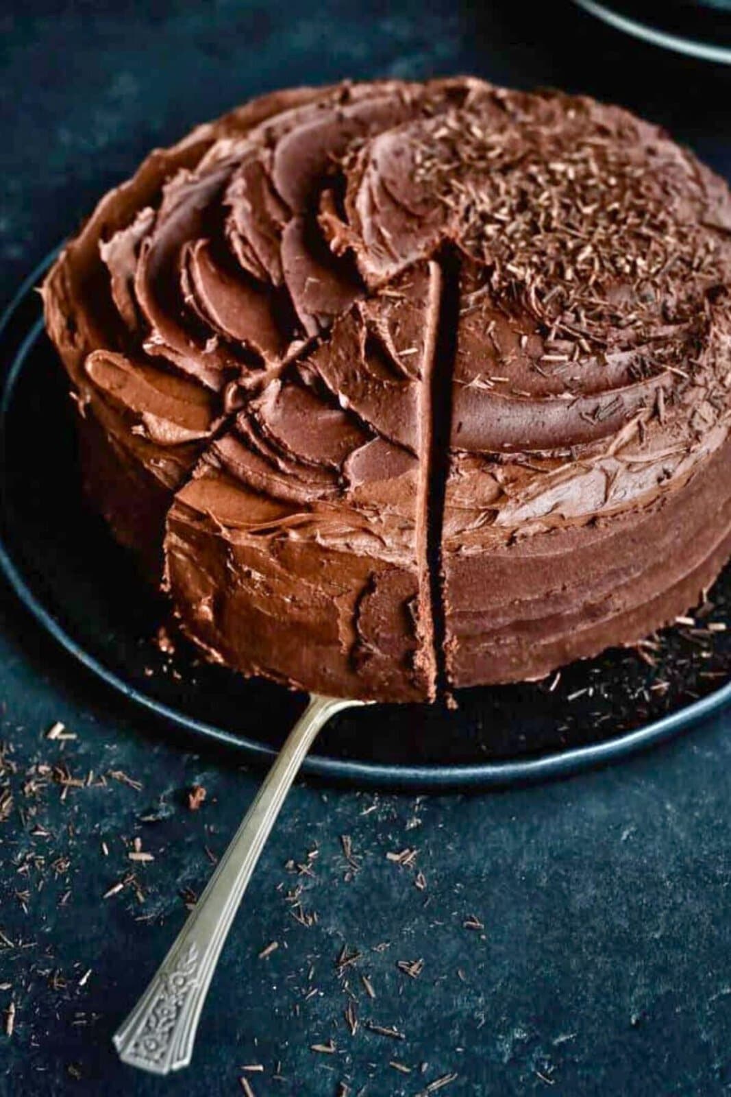 A round Air Fryer Chocolate Cake with swirled chocolate frosting and chocolate shavings on top sits on a black plate. One slice has been cut, and a cake server is beneath it, ready to lift the piece.