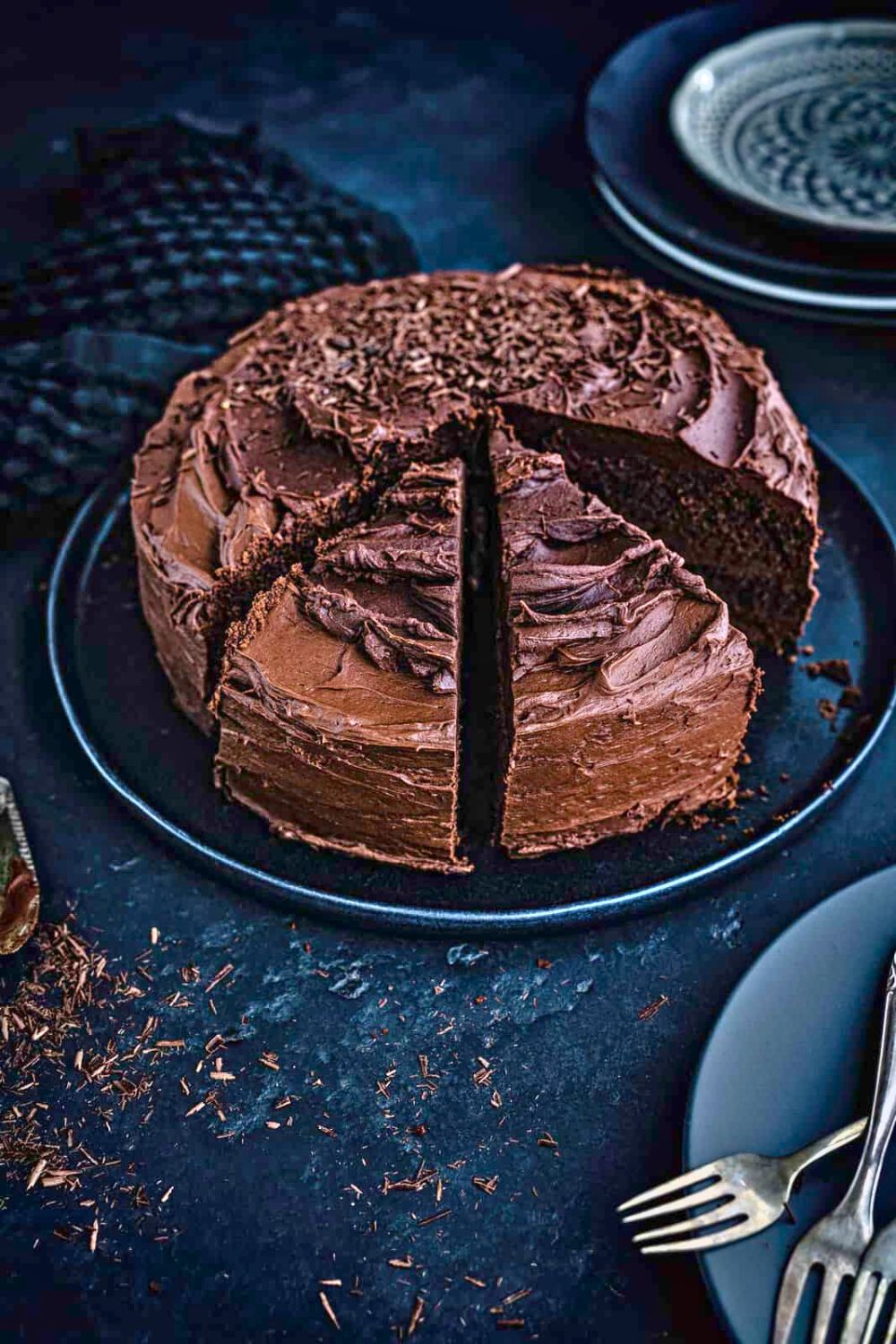 A round Air Fryer Chocolate Cake with rich chocolate frosting, partially sliced, sits on a dark plate. Chocolate shavings decorate the cake, with a fork and plate nearby on a dark, textured surface.