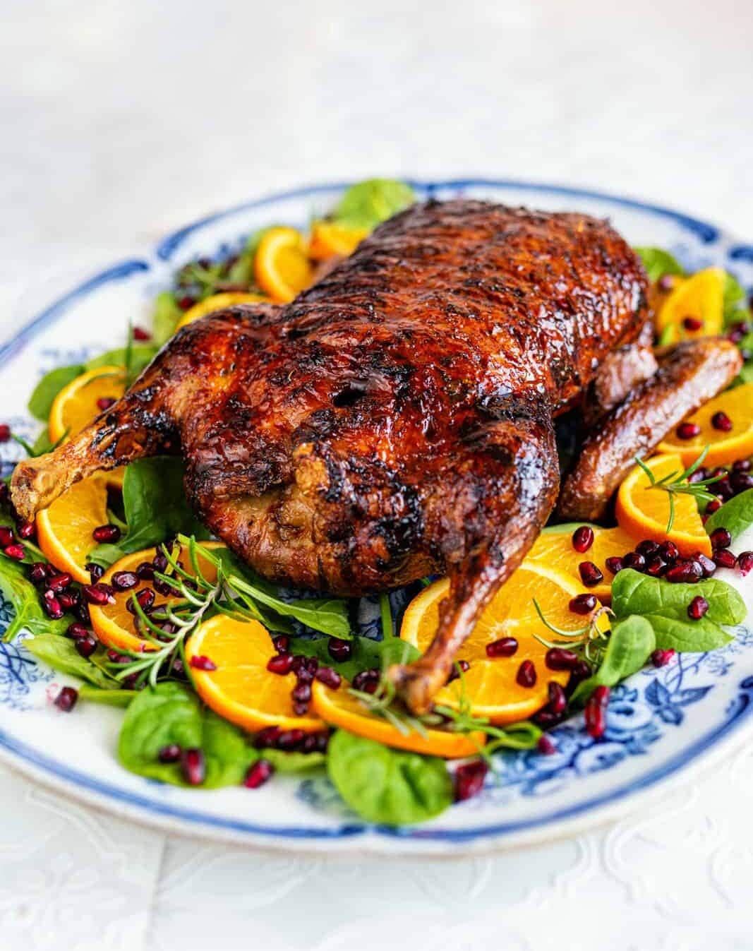 A whole roasted air fryer duck with crispy, golden-brown skin is served on a platter with fresh orange slices, leafy greens, rosemary, and scattered pomegranate seeds.