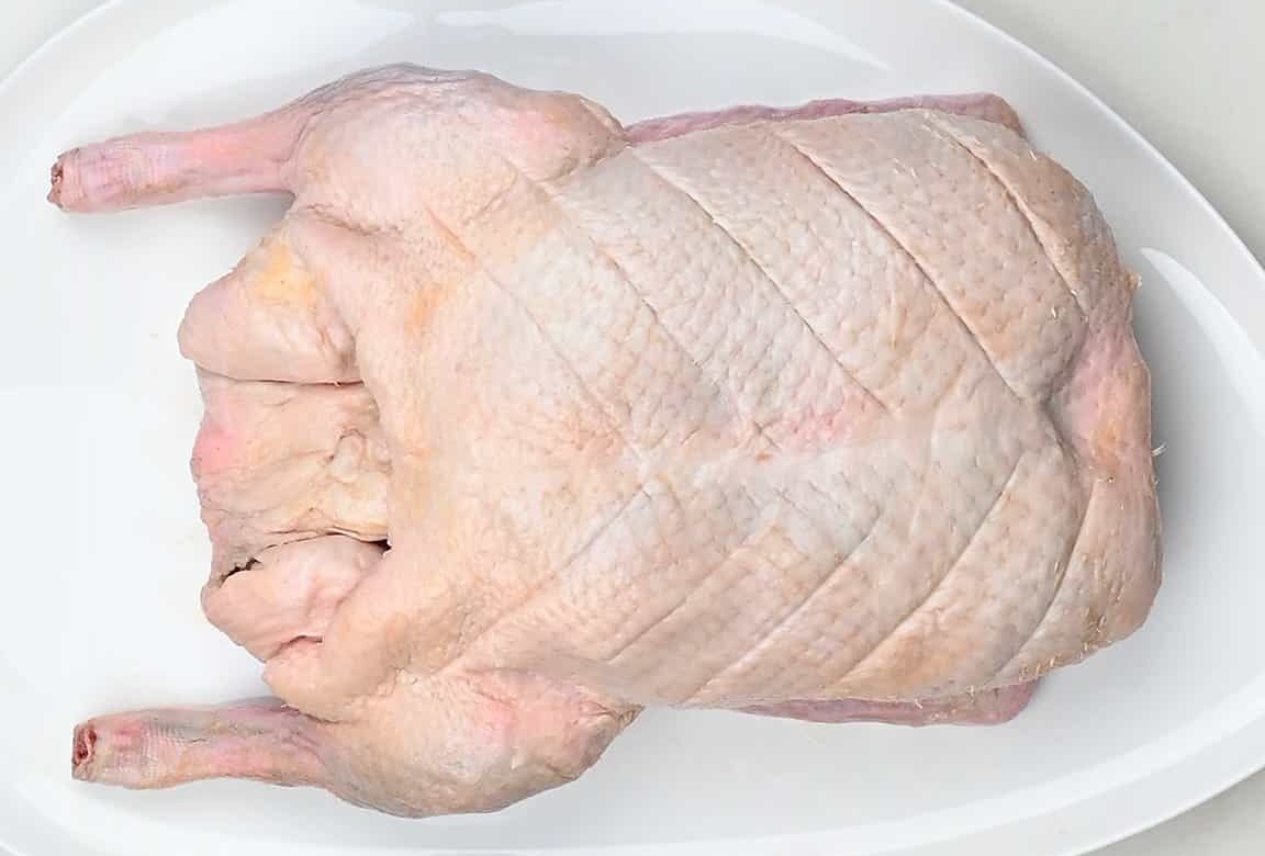 A whole raw duck with scored skin lies on a white oval plate, perfect for preparing Air Fryer Duck at home.