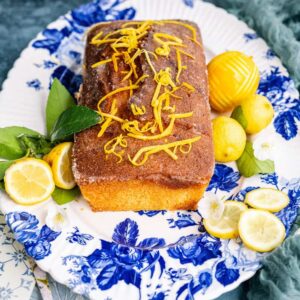 A rectangular air fryer lemon drizzle cake topped with lemon zest sits on a blue and white floral platter, surrounded by whole and sliced lemons, green leaves, and white flowers. A decorative cake server is nearby.