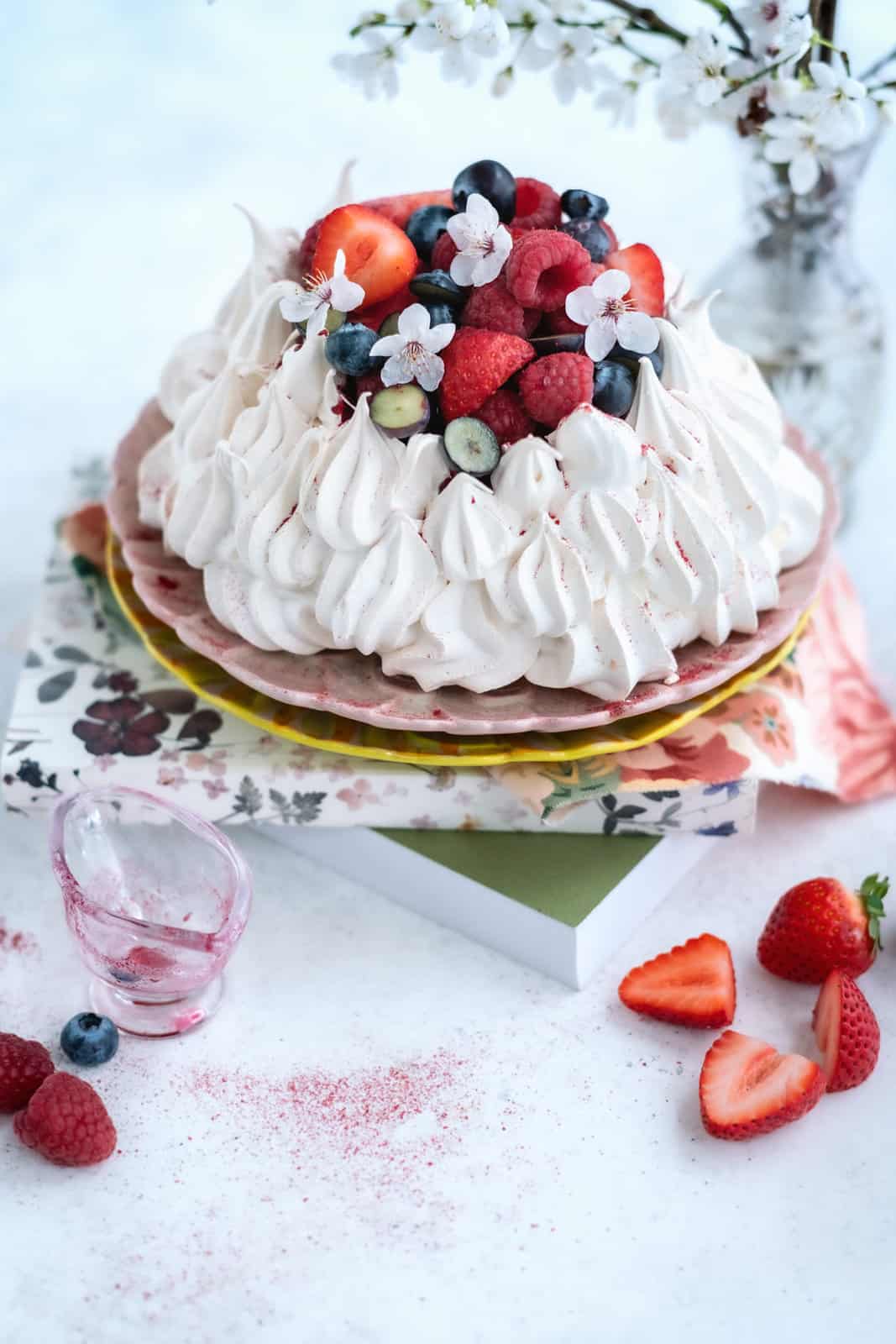 A pavlova topped with whipped cream, strawberries, raspberries, blueberries, and edible flowers sits on stacked floral plates. Fresh berries and a small glass cup are nearby—perfect for serving Air Fryer Pavlova—with white blossoms in the background.