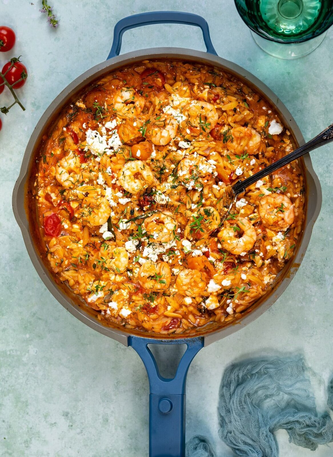 A blue-handled skillet filled with savory shrimp orzo, tomatoes, herbs, and crumbled cheese, with a spoon in the dish. A glass of water, cherry tomatoes, and a blue cloth are nearby on a light surface.