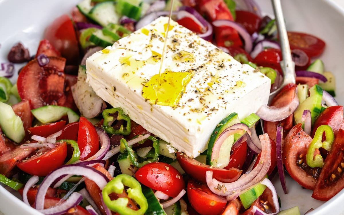 Greek Salad ( Horiatiki Salt) in a white bowl being drizzled with olive oil