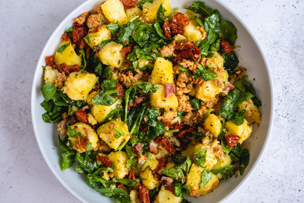 Warm Potato salad with sausage, bacon, sun-dried tomatoes and spinach in a bowl