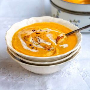 A bowl of creamy Roasted Butternut Squash Soup, garnished with a swirl of cream, croutons, and cracked black pepper, sits on stacked dishes with a spoon, while another pot of soup waits in the background on a white tablecloth.