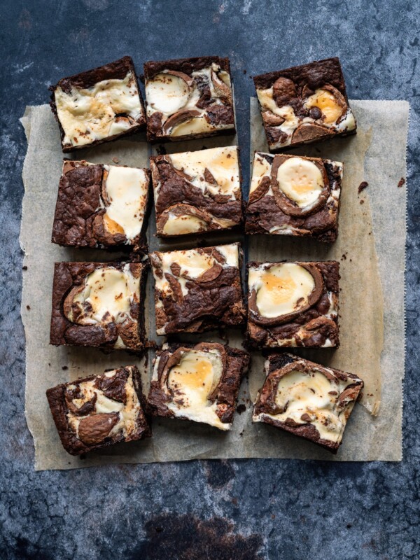 Twelve square air fryer creme egg brownies with swirls of cream cheese are arranged on a sheet of parchment paper atop a dark surface. The brownies have a marbled appearance with dark chocolate and creamy white sections.