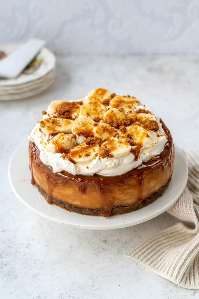 Banoffee cheescake topped with whipped cream, bananas, toffee sauce and biscuit crumbs on a cake stand