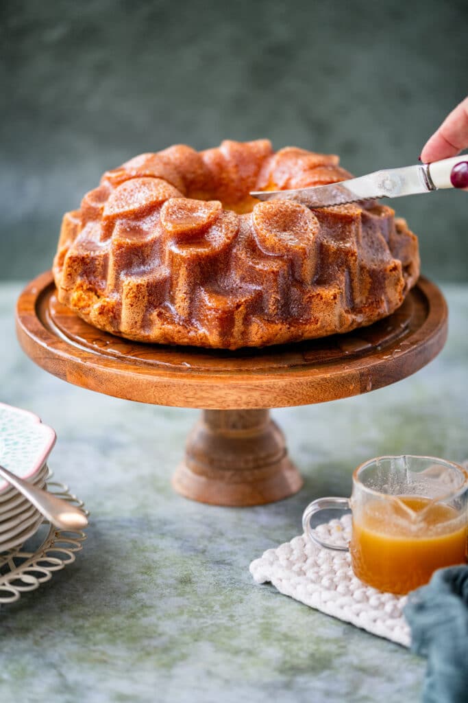 slicing rum cake on a cake stand, jug of rum sauce on the side