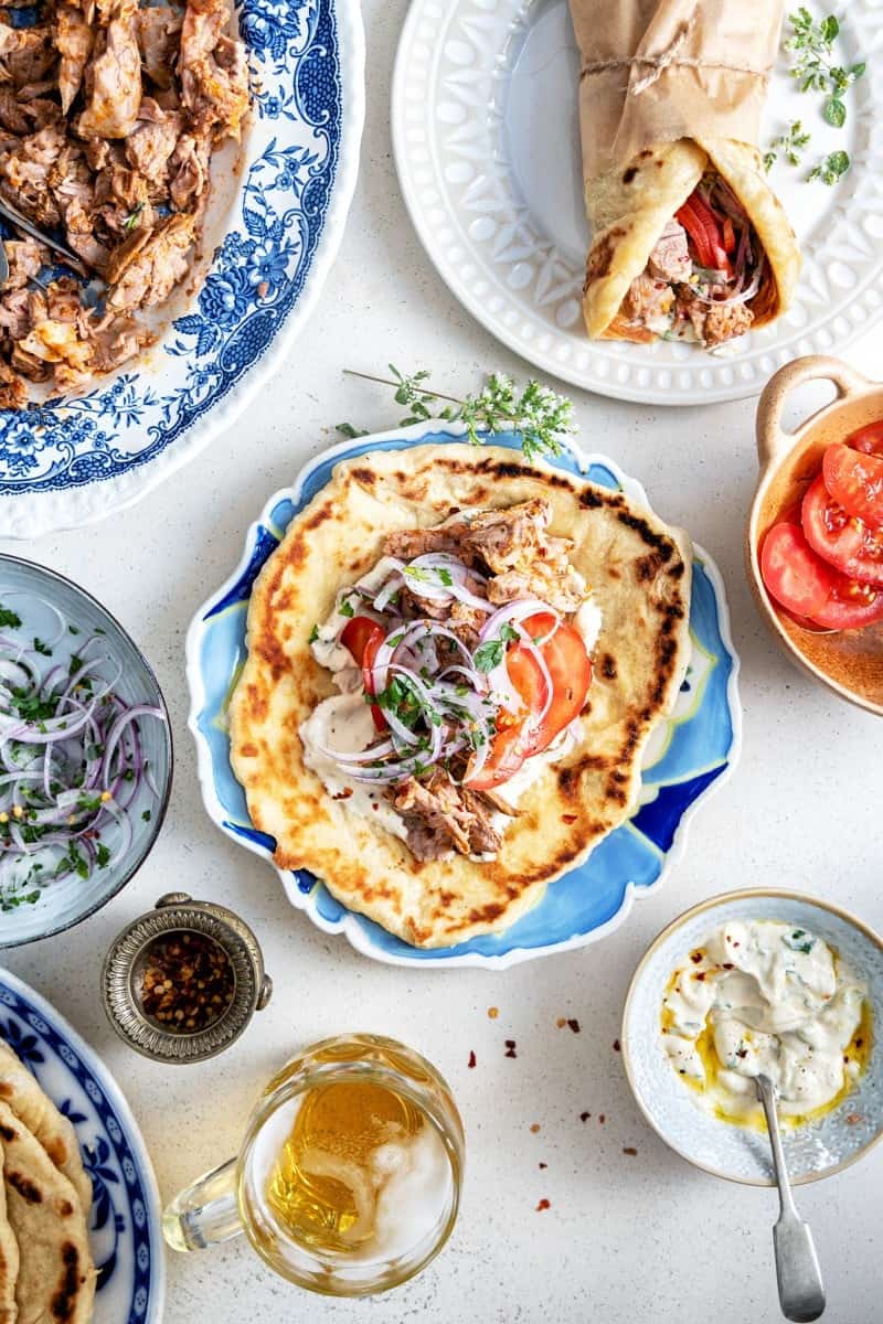 Flat lay of a Mediterranean meal featuring a pita wrap with sliced tomatoes and red onions on a blue plate, surrounded by dishes of lamb gyro meat, a beer glass, sliced tomatoes, and tzatziki sauce on a light surface.