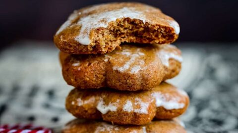 A stack of four iced Lebkuchen gingerbread cookies sits on a decorative surface. The top cookie has a bite taken out of it, while a red and white string rests nearby. The background is dark and blurred.
