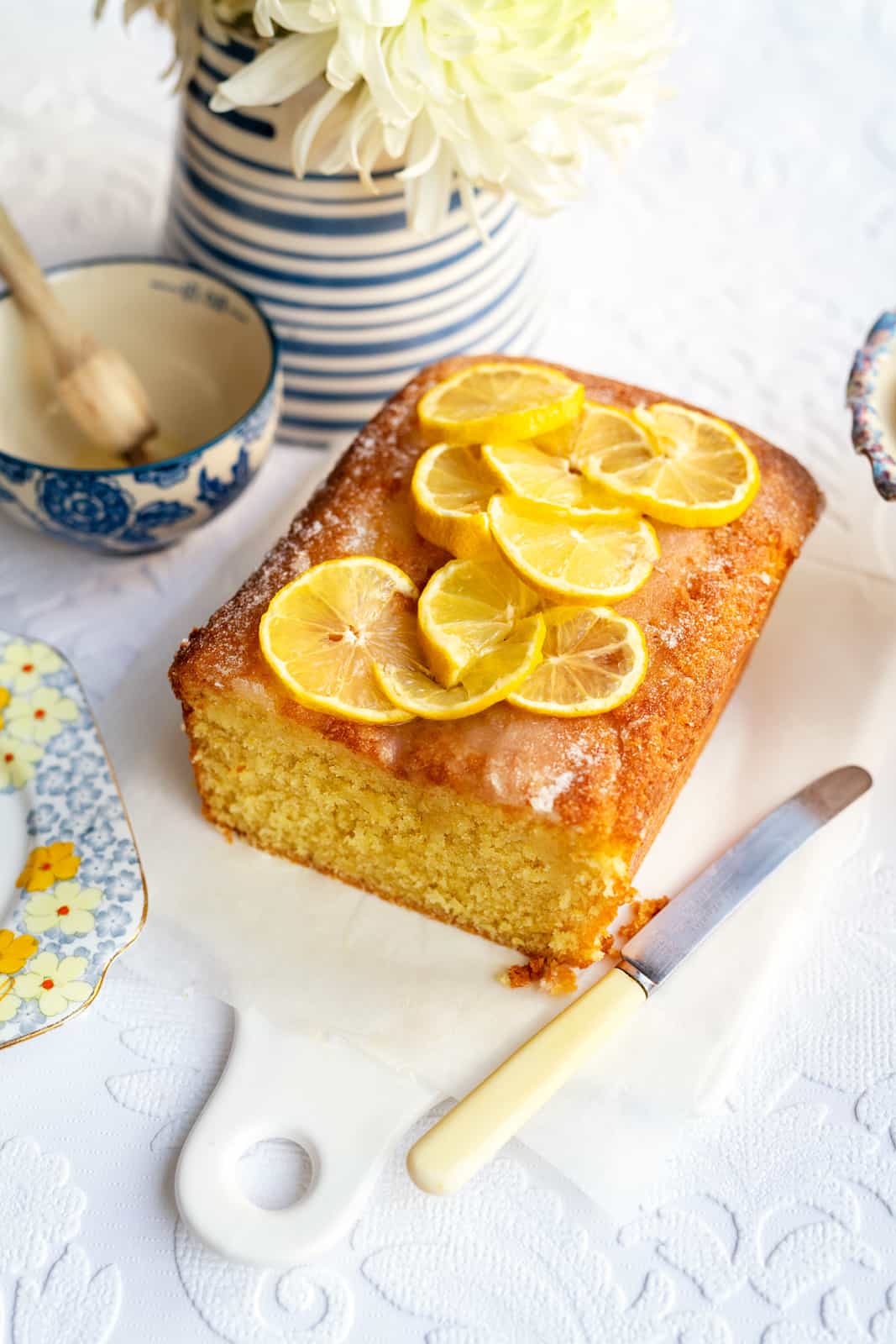 A lemon drizzle cake topped with lemon slices is on a white cutting board. A knife with a yellow handle is next to it. Nearby, a blue and white striped vase holds white flowers, and a floral-patterned cup contains a honey dipper.