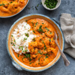 A bowl of creamy shrimp curry, served alongside white rice. The dish is garnished with fresh chopped cilamtro. A small bowl with more herbs and a textured napkin are beside the bowl. A spoon rests in the curry.