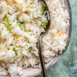 A close-up of a bowl of fluffy coconut rice garnished with thin green herbs, with a metal spoon resting inside the bowl. The background is a textured light green surface.