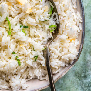 A close-up of a bowl of fluffy coconut rice garnished with thin green herbs, with a metal spoon resting inside the bowl. The background is a textured light green surface.