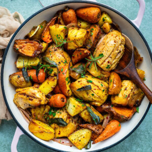 A round white pot with pink handles filled with roasted vegetables, including potatoes, carrots, and herbs, sits on a light green surface. This one pan autumn chicken dinner can be made easily in an air fryer or oven. A wooden spoon rests in the pot.