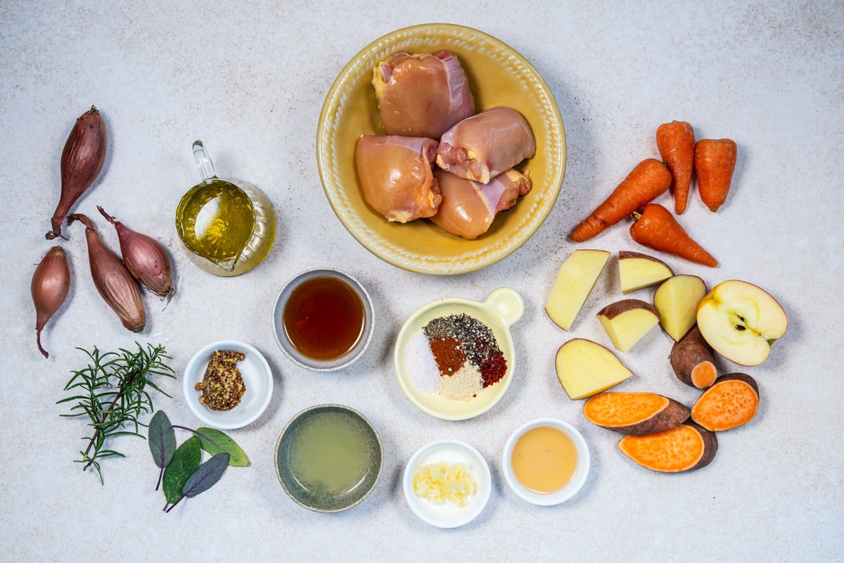 Overhead view of raw chicken thighs in a bowl, surrounded by shallots, olive oil, carrots, apple, sweet potato, yellow potato, herbs, spices, mustard, broth, chopped garlic, lemon juice, and honey on a light surface.