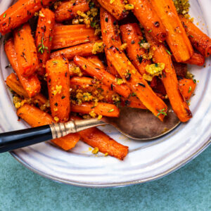A plate of air fryer roasted carrots garnished with herbs and breadcrumbs, served with a spoon on a white dish against a light blue background.