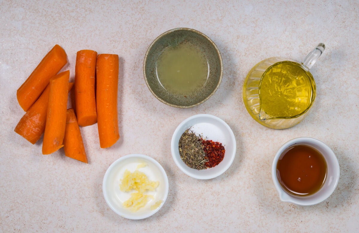 Six large carrot pieces, a bowl of minced garlic, a bowl of mixed dried herbs and red pepper flakes, a bowl of olive oil, a small dish of lemon juice, and a cup of brown liquid are arranged on a light surface.
