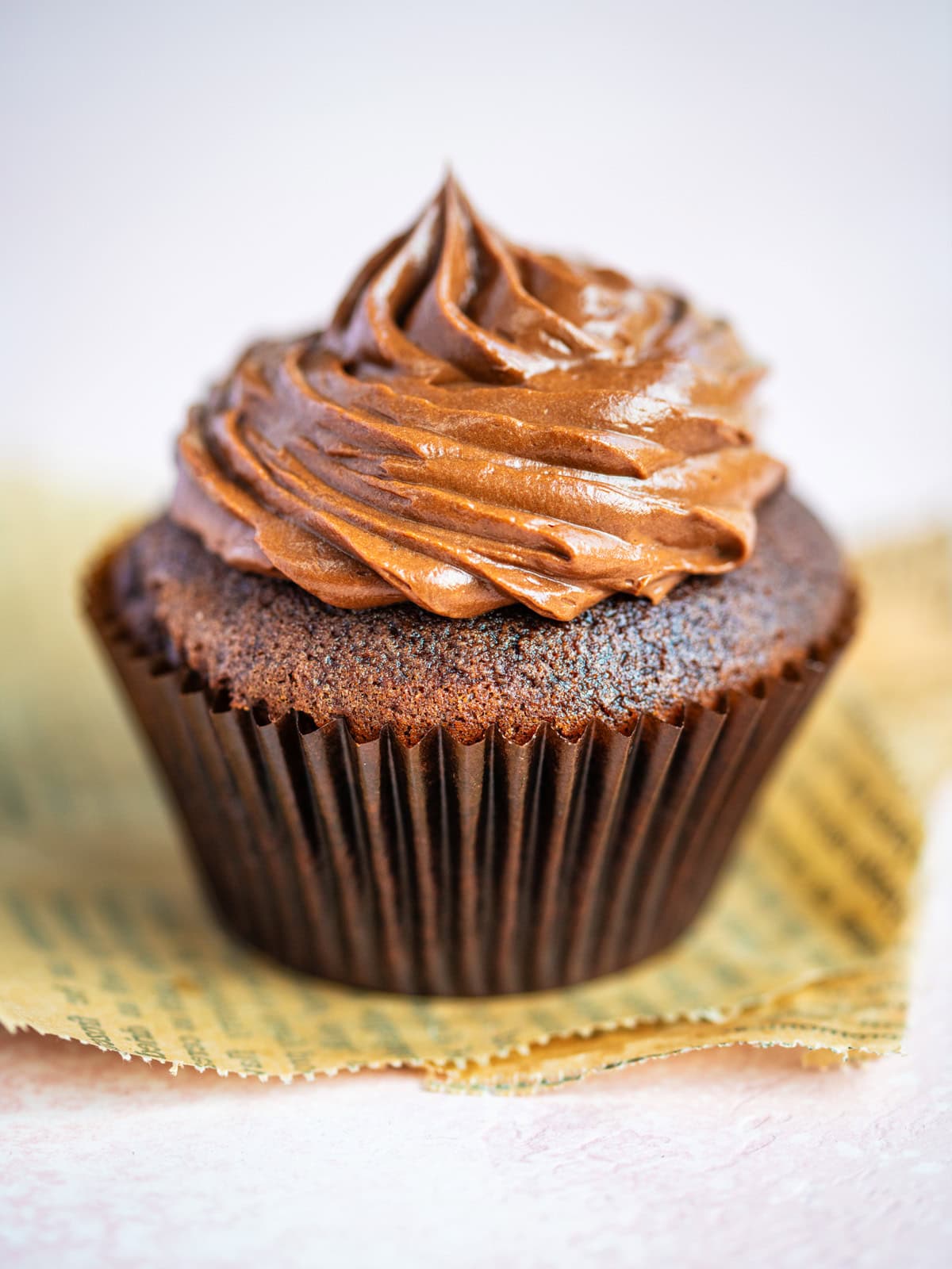 A close-up of a chocolate cupcake in a brown paper liner, topped with swirled chocolate frosting, sits on a piece of parchment paper against a light background.